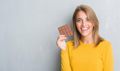 Beautiful young woman over grunge grey wall eating chocolate bar with a happy face standing and smiling with a confident smile showing teeth