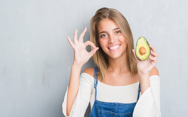 Beautiful young woman over grunge grey wall eating avocado doing ok sign with fingers, excellent symbol
