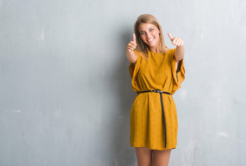 Beautiful young woman standing over grunge grey wall wearing a dress approving doing positive gesture with hand, thumbs up smiling and happy for success. Looking at the camera, winner gesture.