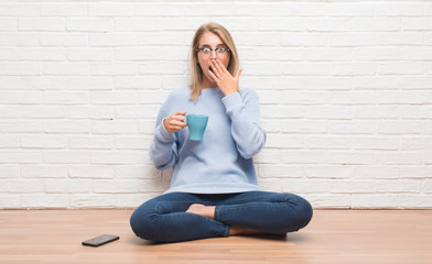 Fototapeta na wymiar Beautiful young woman sitting on the floor driking cup of coffee at home cover mouth with hand shocked with shame for mistake, expression of fear, scared in silence, secret concept