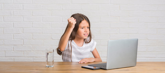 Young hispanic kid sitting on the table using computer laptop annoyed and frustrated shouting with...