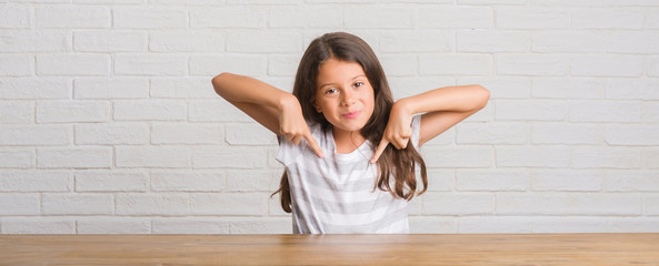 Young hispanic kid sitting on the table at home looking confident with smile on face, pointing...