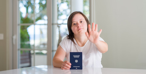 Obraz premium Down syndrome woman at home holding german passport with open hand doing stop sign with serious and confident expression, defense gesture