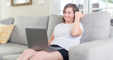 Down syndrome woman at home using computer laptop pointing and showing with thumb up to the side with happy face smiling
