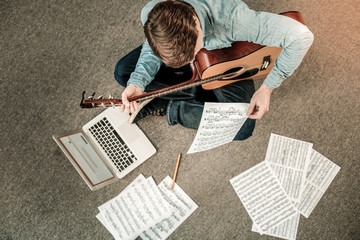 Young musician. Happy positive young man sitting on the floor and looking at the notes while...