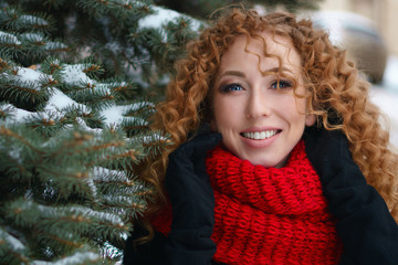 Girl with red hair in a fur coat and red knitted scarf walks outdoors in winter. She stands of spruce in snow.