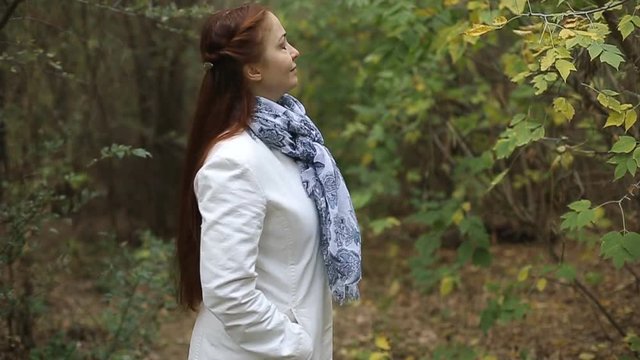 red-haired Caucasian middle-aged woman in a white coat walks on a cloudy day in the autumn park
