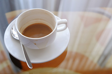 White ceramic cup of almost empty coffee with coaster and metal spoon, on the transparent glass table with the reflection of curtain on the surface
