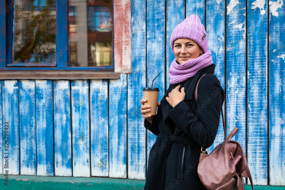 Wall mural A young woman in a pink knitted hat, black coat and backpack  enjoys city, poses and drink coffee to go around blue wall. The concept of livestyle and outdoor recreation in autumn