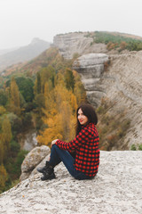 Traveler woman wearing red plaid shirt and jeans sitting on mountain and looking at autumn forest. Hiking in autumn. Wanderlust concept. Cloudy weather, fall colors, autumn landscape.