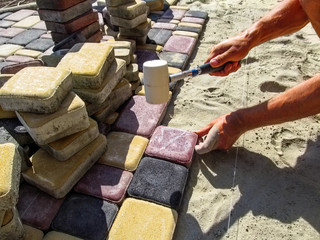 A worker in the process of laying multi-colored paving slabs. A man installs paving bricks on a sand-cement mixture using a rubber mallet