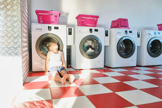 Cute little boy in the Laundromat is tired, sitting on the floor and waiting for the Laundry to finish. Room with laundry machines, Sunny summer day.