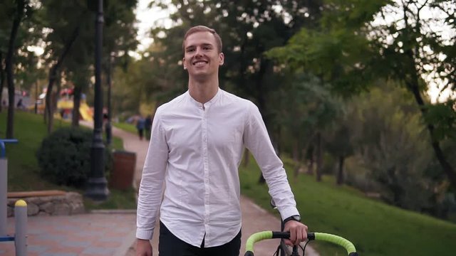 Caucasian smiling young man in white shirt walking with bicycle on the street in town. Rolling his trekking bike while walking by park. Front view