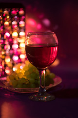 A glass of red grape wine, a plate with bunches of grapes in a cozy atmosphere in the evening against the backdrop of romantic lights with a bokeh effect