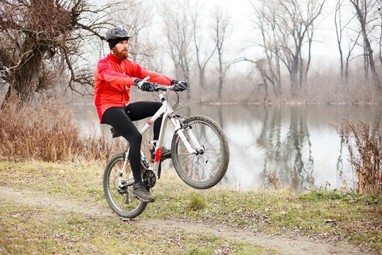 Determined young bearded man doing a wheelie on a mountain bike while riding by the river on a foggy autumn or winter day