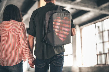 Heart on backpack. Young man wearing casual clothes and carrying backpack while holding hands with his dark haired girlfriend