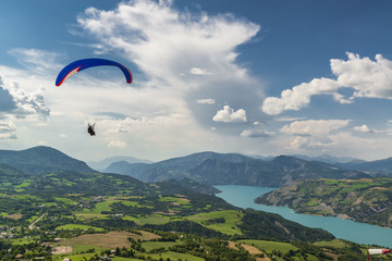 Bright paraglider tandem wing fly over beautiful mountain valley with green hills and blue lake.