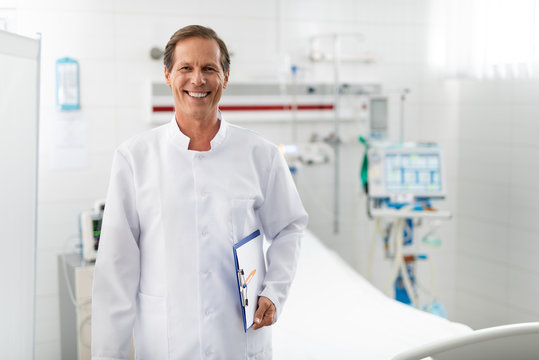 Waist up portrait of middle aged man in white lab coat holding clipboard while looking at camera with wide smile. Medical equipment and bed on blurred background