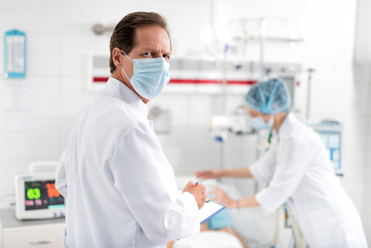 Waist up portrait of middle aged man in white lab coat holding clipboard and pen. Female assistant putting oxygen mask on patient on blurred background