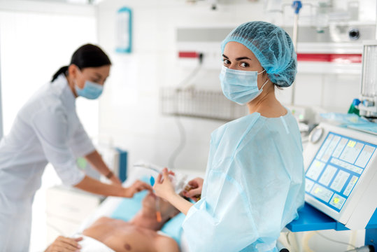 Ready To Help. Waist Up Portrait Of Female Medical Worker In Protective Mask And Cap Looking At Camera While Young Doctor Preparing Patient For Mechanical Ventilation