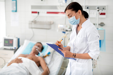 Side view portrait of young lady in protective face mask holding clipboard and noting information about condition of middle aged man. Gentleman on breathing machine lying in hospital bed on blurred
