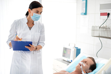 Waist up portrait of young lady in protective mask and white lab coat noting diagnosis of middle aged man. Sleeping gentleman lying in bed with endotracheal tube in mouth