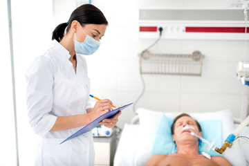 Side view portrait of young woman in protective mask holding clipboard and filling up medical form. Middle aged man on mechanical ventilation on blurred background