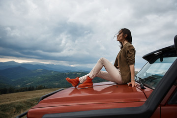 Delighted young woman resting on the car hood while spending her weekend in the mountains