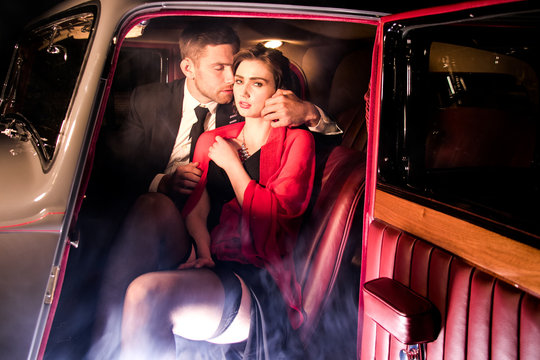Good looking sexy couple, handsome man in suit, beautiful woman in red dress, embrace passionately in vintage car