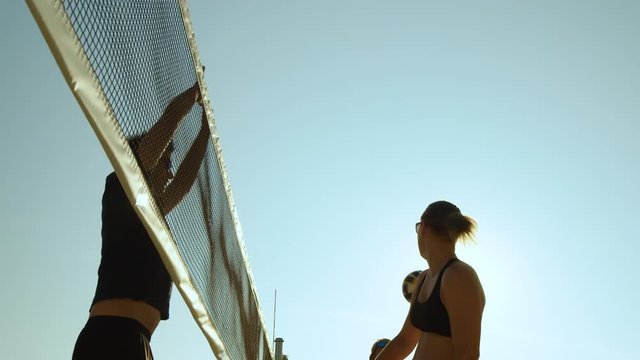 SLOW MOTION, CLOSE UP, LOW ANGLE, LENS FLARE: Tall man blocks the ball spiked by athletic female volleyball player. Fit women and man enjoying summer vacation playing beach volleyball in the hot sun.