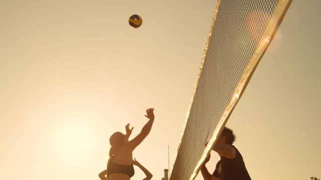 SLOW MOTION, LOW ANGLE, CLOSE UP, LENS FLARE: Fit young people having fun playing beach volleyball at sunset. Young Caucasian girl spikes the ball over the net and man blocks it. Cool summer sport.