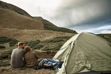 Fototapeta premium Cuddling near tent. Peaceful young couple sitting near the tent and looking relaxed while enjoying beautiful landscape