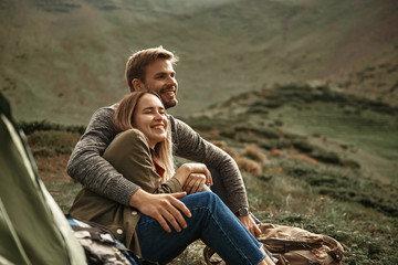 Warm hug. Calm kind loving man sitting near the tent in the mountains and smiling while hugging his...