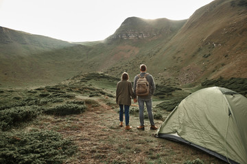 Fototapeta premium Couple near tent. Peaceful relaxed young man and woman standing near the tent and holding hands while looking at the landscape