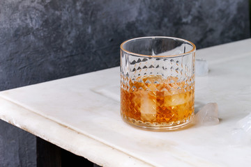 Glass of scotch whiskey on ice standing on white marble table with grey wall at background. Alcohol drink.