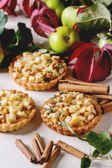 Homemade sweet apple shortbread tartlets with cinnamon sticks, walnuts, apples red leaves branches above on white marble table. Autumn baking.