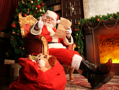 Portrait of happy Santa Claus sitting at his room at home near Christmas tree and reading Christmas letter or wish list.