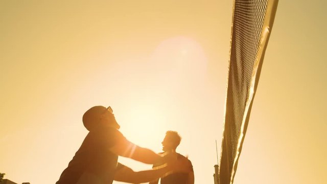 SLOW MOTION, CLOSE UP, LOW ANGLE, LENS FLARE: Cheerful male volleyball player spikes ball into the net at sunset. Guys on active summer holiday having fun playing beach volleyball on a sunny morning.
