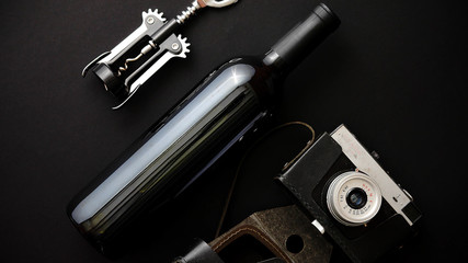 Red wine bottle, corkscrew and old photo camera on black matte background. Top view with copy space