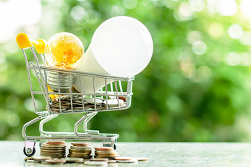 Led lamp and glowing light bulb in mini shopping cart or trolley  with money coins against blurred...