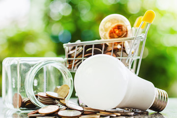 Fototapeta na wymiar Glowing light bulb and coins in mini shopping cart or trolley with LED lamp and the glass jar of money against blurred natural green background for finance, saving energy and environment concept