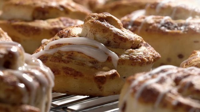 Cinnamon bun on cooling rack with icing being dripped over the cinnamon bun in slow motion. 