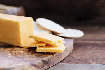 Block of cheddar cheese and slices over a rustic background.. Extreme shallow depth of field with selective focus on cheese. Water crackers in  blurred background.
