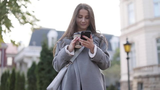 beautiful woman in brown coat goes near in the old city, communicates via smartphone (phablet). Steady cam shot, slow motion.autumn look