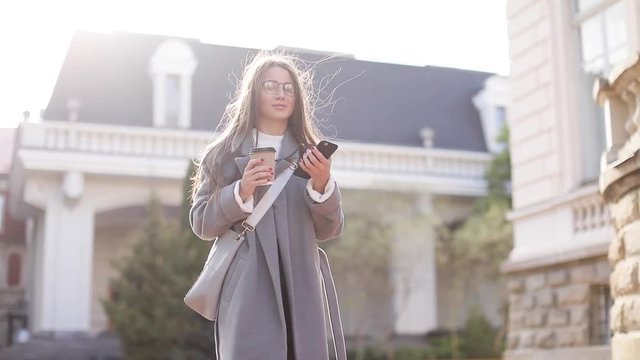 beautiful woman in brown coat goes near in the old city, communicates via smartphone (phablet). Steady cam shot, slow motion.autumn look