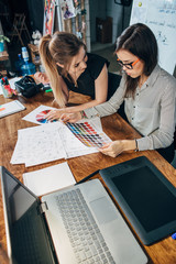 Young female graphic designers sitting at desk with laptop, color charts and some sketches...