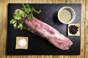 pork tenderloin next to salt, black pepper, a bowl of oil and parsley on a black slate plate on a wooden table
