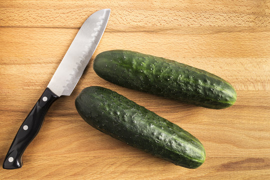 two whole green cucumbers next to a knife on a wooden table
