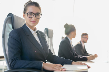 Serious confident young female business student in glasses sitting at table in row of colleagues and looking at camera while listening to seminar in conference room.
