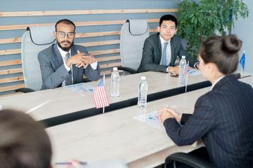 Serious pensive multi-ethnic diplomats in formal suits sitting at conference table with national...